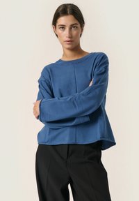 Blå langærmet sweatshirt med rund halsudsk�æring, der har en afslappet pasform og blød tekstur. Matchet med sorte bukser uden synlige mønstre.