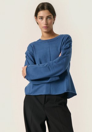 Blå langærmet sweatshirt med rund halsudskæring, der har en afslappet pasform og blød tekstur. Matchet med sorte bukser uden synlige mønstre.
