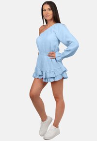 Lichtblauwe, gestructureerde jurk met één schouder, lange mouw en gerafelde zoom, gecombineerd met witte sneakers.