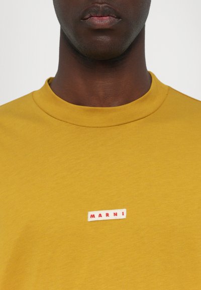 Tricou scurt, de culoare galben muștar, cu guler rotund; prezintă o etichetă albă cu text roșu care spune "MARNI" pe față. Textură netedă și moale.