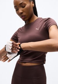 Αθλητική γυναίκα που φοράει καφετί cropped top Puma και κολάν, προσαρμόζει λευκά γάντια γυμναστικής χωρίς δάχτυλα μπροστά σε απλό λευκό φόντο.