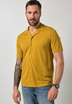 Mann mit Bart und tätowiertem Arm trägt ein senfgelbes kurzärmeliges Hemd und blaue Jeans, steht vor einem schlichten Hintergrund.