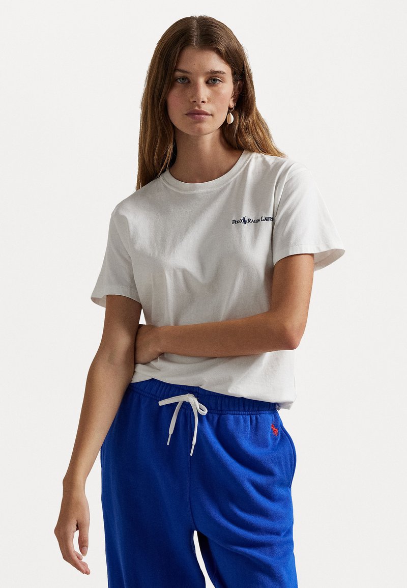 Balta medvilninė marškinė su apvalia iškirpte ir trumpomis rankovėmis, ant kurių siuvinėtas mažas logotipas. Derinama su mėlynomis sportinėmis kelnėmis su raišteliu.