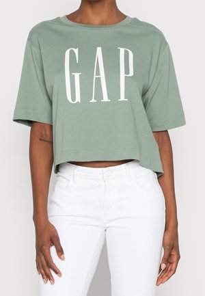 Osoba nosząca przyciętą bluzkę w kolorze szałwii z dużym białym napisem „GAP” oraz białe spodnie z wysokim stanem, stojąca na jednolitym tle.