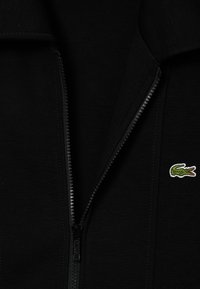 Sudadera negra con cremallera fabricada en un tejido texturizado, que presenta un logotipo de cocodrilo bordado en verde en el lado izquierdo.