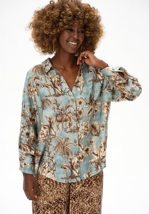 Uśmiechnięta kobieta o kręconych włosach, ubrana w niebieską koszulę zapinaną na guziki z nadrukami zwierząt i liści, w połączeniu z brązowymi spodniami w panterkę.