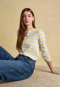 Tânără cu părul lung și castaniu stând pe scaun, purtând un pulover alb cu dungi galbene și albastre și blugi albaștri, pe un fundal bej.