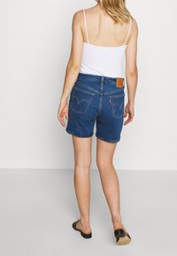 Pantalones cortos de mezclilla en azul oscuro con un corte clásico, cinco bolsillos y un parche de marca de cuero en la parte trasera. Presenta detalles de costura sutiles.