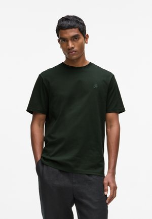 Fiatal férfi áll, kezét a zsebében tartva, sötétzöld rövid ujjú pólót és sötét nadrágot visel, egyszínű háttér előtt.