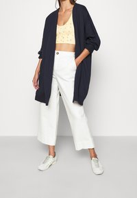 Navy langer Cardigan über einem hellgelben blumigen Crop-Top, kombiniert mit weißen weit geschnittenen Hosen und wei�ßen Sneakers mit grünen Akzenten.