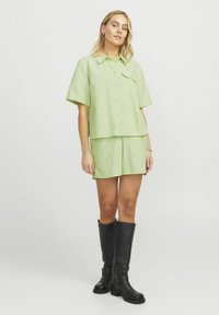 Camicia verde chiaro a maniche corte con tasca, abbinata a pantaloni corti coordinati. Il modello indossa stivali neri alti. Tessuto liscio, motivo sottile.