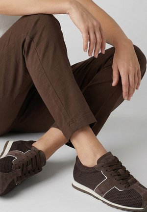 Personne assise les jambes croisées portant un pantalon marron et des baskets en mesh marron foncé avec des accents blancs sur un sol gris clair.