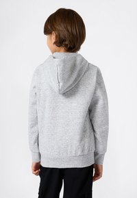 Niño con cabello corto castaño, vistiendo una sudadera gris claro y pantalones negros, de espaldas contra un fondo blanco liso.