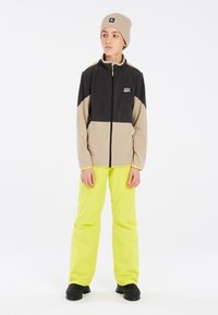 Fleecejas met zwarte en beige panelen, voorrits en logopatch. Gecombineerd met felgele broek en zwarte winterlaarzen.