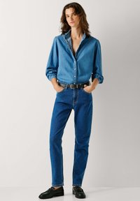 Džínová košile s dlouhými rukávy a zapínáním na knoflíky, spárovaná s modrými džíny a černým pásem. Celý outfit doplňují ploché černé boty.