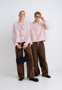 Δύο άτομα φοράνε ροζ ανοιχτού χρώματος, ριμπ βαμβακερά κοντά πουκάμισα με κουμπιά και δεσίματα, συνδυασμένα με σκούρα καφέ φαρδιά παντελόνια και μαύρα παπούτσια.