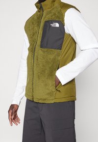 Olivgrön väst med texturerad, fluffig front och svart ficka med en vit logotyp. ärmlös, med dragkedja och sidfickor.