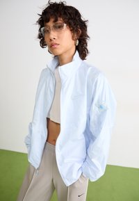 Persona che indossa una giacca a zip di colore azzurro chiaro sopra una maglietta corta bianca e pantaloni beige Nike, in piedi con le mani in tasca contro uno sfondo neutro.