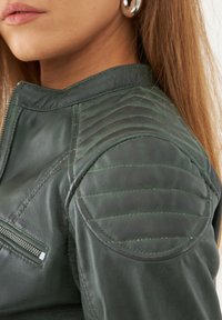 Tummanvihreä nahkatakki, jossa on tikkaukset hartioissa ja vetoketjullinen tasku. Sujuva pinta, istuva malli ja korkea kaulus.