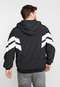 Sort hættejakke med hvide striber på ærmerne, fremstillet af letvægtsmateriale. Har en afslappet pasform og elastiske detaljer ved håndleddene.