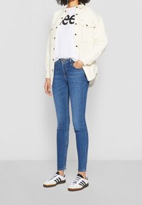 Persona con camisa de botón crema sobre una camiseta blanca con logotipo, jeans azules ajustados y zapatillas blancas con rayas negras, de pie contra una pared blanca.