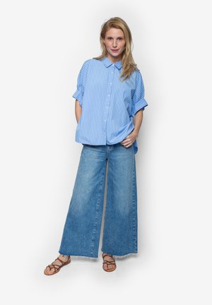 Frau trägt ein blau gestreiftes Hemd mit Knopfleiste, weit geschnittene Jeans und braune Riemchensandalen, steht vor einem weißen Hintergrund.