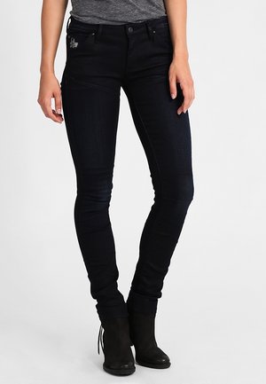 Femme portant un jean noir slim et des bottines noires, debout avec les mains détendues le long du corps.