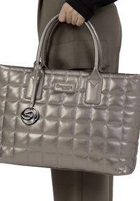 Sac fourre-tout matelassé argent métallisé avec double poignées, doté d'un charme décoratif en forme de logo et d'une étiquette de marque rectangulaire. Texture en cuir lisse.