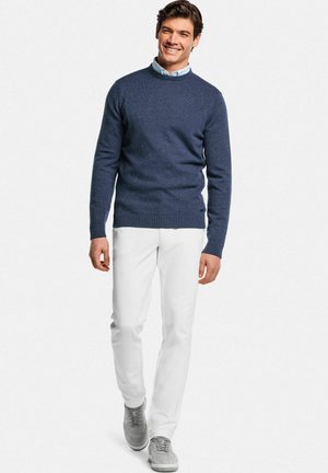 Lachende man die vooruit loopt, gekleed in een blauw trui met ronde hals, lichtblauw overhemd met kraag, witte broek en grijze sneakers op een witte achtergrond.