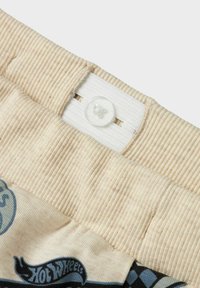 Beige ribattu vyötärönauha, jossa on valkoinen joustava insert ja kirkas muovipainike. Alla kuviol fabric sinisillä kuvioilla ja tekstillä.