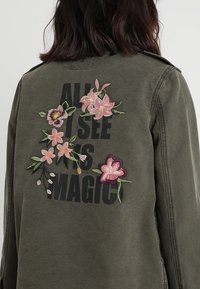 Oliwkowa zielona kurtka z kwiatowym haftem w kolorach różowym i fioletowym na plecach, z napisem "ALL I SEE IS MAGIC" w kolorze czarnym.