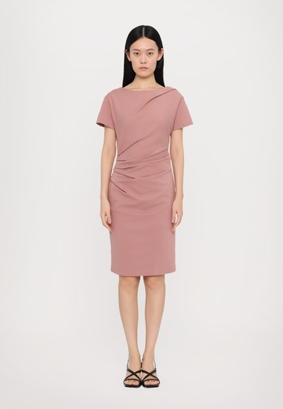 Knieumspielendes, figurbetontes Kleid aus blassrosa Stoff mit einem drapierten Ausschnitt und dezentem seitlichen Raffung. Kombiniert mit schwarzen Riemchensandalen.
