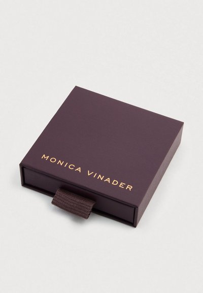 Cutie mov închis cu finisaj mat, cu logo-ul "MONICA VINADER" embosat în aur și o mică buclă textilă maro.