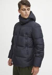 Pufferjakke i mørk marineblå, kviltet design med høyt krage og avtagbar hette. Har en glidelåslomme på brystet. Myk, glatt tekstur.