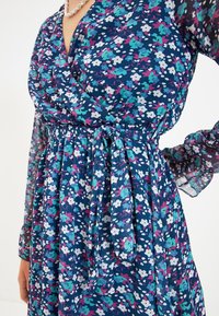 Kukallinen vaaterengas tummansinisenä, jossa on vaaleanpunaisia, teal- ja valkoisia kuvioita. Ominaisuuksiin kuuluu sidottu vyötärö ja läpinäkyvät pitkät hihat, valmistettu kevyestä kankaasta.