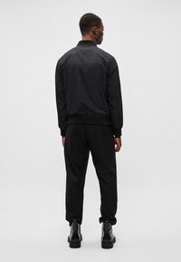 Schwarze Bomberjacke mit gerippten Bündchen und Saum, getragen über passenden schwarzen Hosen und massiven schwarzen Stiefeln, von hinten betrachtet.