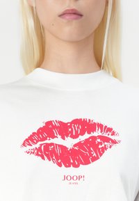 Weißes T-Shirt mit einem roten Lippenabdruck und dem Text "JOOP! JEANS" darunter. Einfaches Design mit Rundhalsausschnitt und glatter Textur.