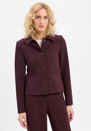 Woman with curly blonde hair wears a tailored dark purple buttoned blazer and matching pants against a plain white background.