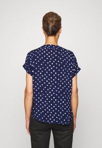 Camisa azul de manga curta com estampa de losangos brancos, corte descontraído, decote redondo e textura suave, vestida por um modelo com calças pretas.