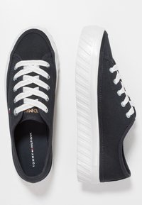 Baskets noires en toile avec des lacets et des semelles blanches. Présentant un logo TOMMY HILFIGER sur la languette et un motif sur le côté de la semelle.