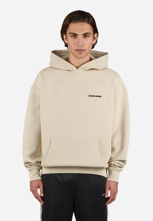 Beige Kapuzenpullover mit einem strukturierten Muster, Fronttasche und schwarzem Logo auf der Brust. Kombiniert mit schwarzen Hosen mit weißen Akzenten.