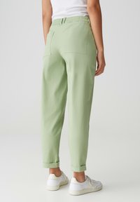 Pantalons vert clair avec une taille élastique, jambes effilées et ourlets roulés. Deux poches arrière et une texture douce. Portés avec des baskets blanches.