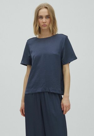 Kobieta o blond włosach, ubrana w luźny granatowy krótki rękaw i pasujące szerokie spodnie na jednolitym tle.