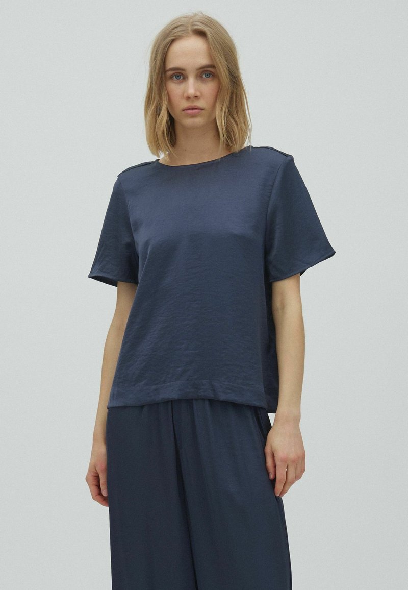 Femme aux cheveux blonds portant un top ample bleu marine à manches courtes et un pantalon large assorti, sur un fond uni.