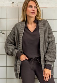 Gebreide grijze cardigan met stevige ribbelstructuur, oversized mouwen en een open voorkant, gedragen over een donkerbruine top. Glad materiaal met een casual pasvorm.