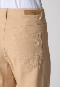 I pantaloni beige vengono mostrati da dietro, evidenziando l'etichetta U.S. Polo Assn. e un piccolo logo ricamato sulla tasca destra.