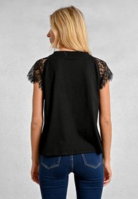 Top nero a maniche corte con dettagli in pizzo sulle spalle, dalla vestibilità rilassata e orlo dritto. Indossato con jeans blu aderenti.