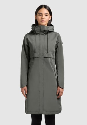 Femeie purtând o haină lungă de iarnă gri, cu glugă, fermoar frontal și buzunare, stând pe un fundal simplu gri deschis.