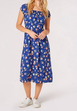 Femme portant une robe midi bleue à imprimé floral rose et blanc, manches courtes, encolure froncée, associée à des baskets blanches.