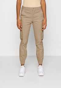 Pantalons cargo beiges avec des poignets élastiques, dotés de poches latérales et d'une texture lisse. Associés à des baskets blanches.